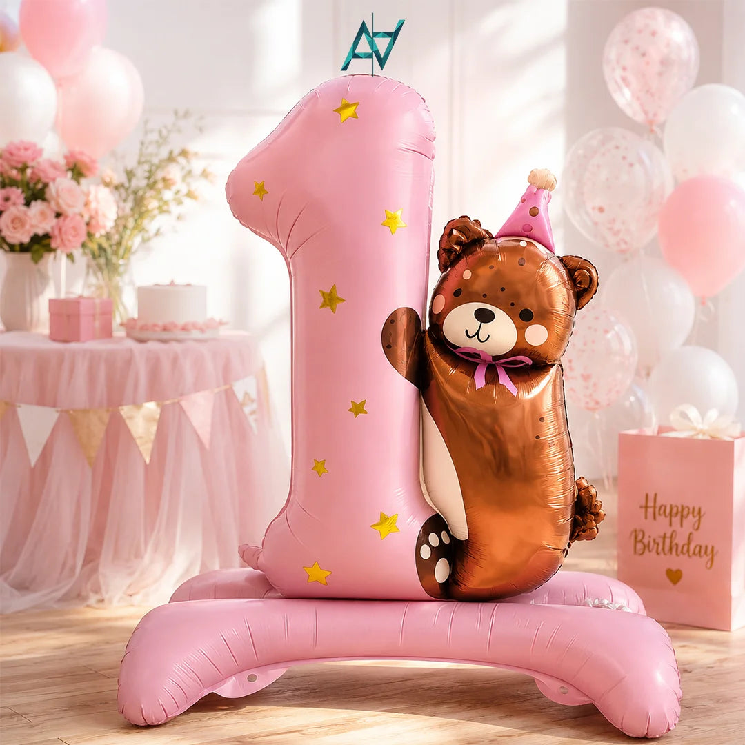 A pink foil balloon shaped like the number 1 stands on a matching base, with a metallic brown teddy bear balloon leaning against it. The scene is a decorated birthday setup with pink balloons, a cake table, and a pink 'Happy Birthday' gift box.